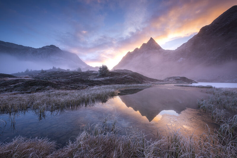 Bergsee in den Schweizer Alpen am frühen Herbstmorgen mit Raureif, Spiegelung, tiefem Nebel und warmem Licht hinter den Bergen.
