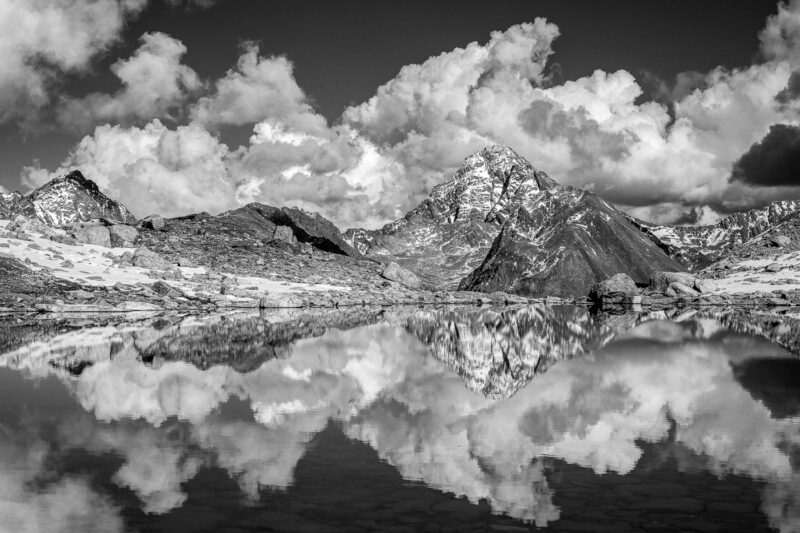 Schwarzweissfotografie eines Berges in den Schweizer Alpen mit mächtigen Quellwolken und Spiegelung in einem Bergsee.