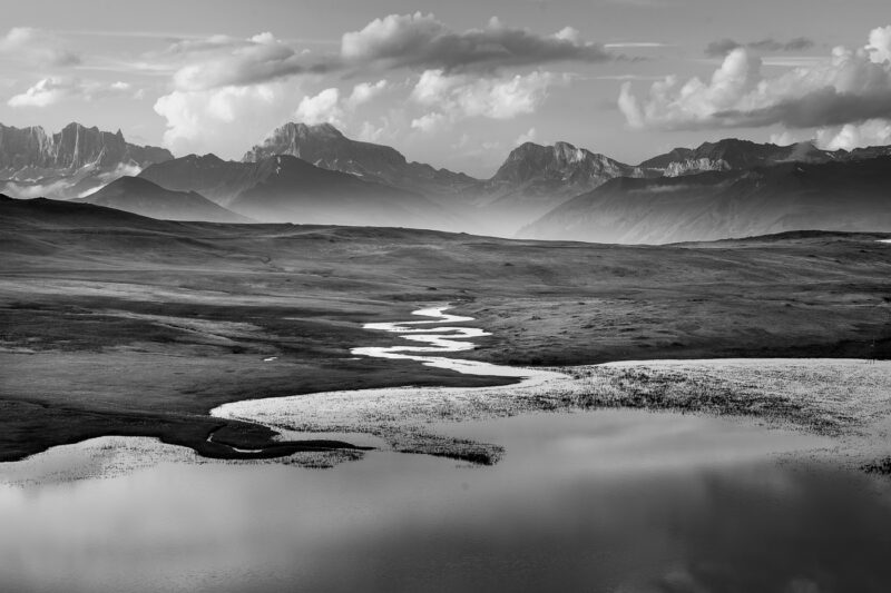 Schwarzweissfotografie einer Schweizer Berglandschaft mit einem geschwungenen Bach, Bergsee und gezackten Gipfeln im Hintergrund.