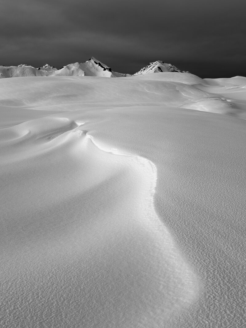 Schwarzweissfotografie einer winterlichen Berglandschaft mit einer geschwungenen, vom Wind geformten Schneekante, die zu den Gipfeln führt.